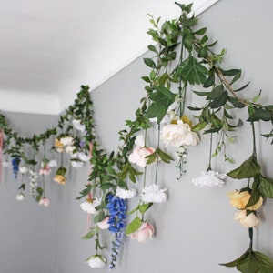 Pu&ograve; includere: Una ghirlanda di fiori e verde artificiali &egrave; appesa a una parete grigia. La ghirlanda presenta fiori bianchi, gialli, rosa e blu, intervallati da foglie verdi e viti. La composizione &egrave; decorativa e aggiunge un tocco naturale.