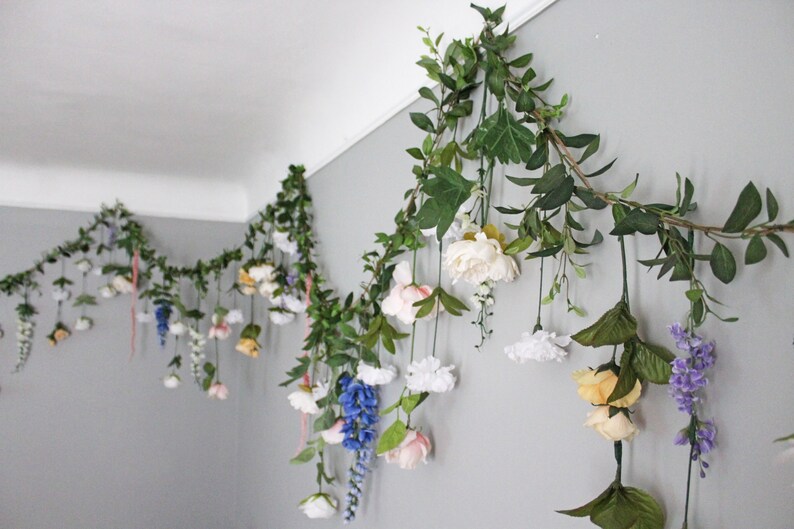 Pu&ograve; includere: Una ghirlanda di fiori e verde artificiali &egrave; appesa a una parete grigia. La ghirlanda presenta fiori bianchi, gialli, rosa e blu, intervallati da foglie verdi e viti. La composizione &egrave; decorativa e aggiunge un tocco naturale.