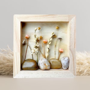 May include: A square wooden frame with a glass front, showcasing a miniature dried flower arrangement with small pink and white flowers, dried grasses, and brown stones.