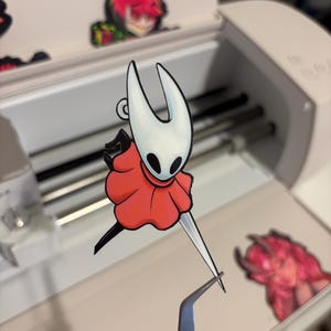 May include: A white and red cartoon character sticker with a long, thin blade. The sticker is being held by tweezers. Other cartoon stickers are visible in the background.