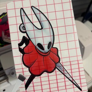 May include: A vinyl sticker featuring a cartoon character with a white head, black eyes, and a red cape. The character is holding a long, silver blade. The sticker is on a red grid background.