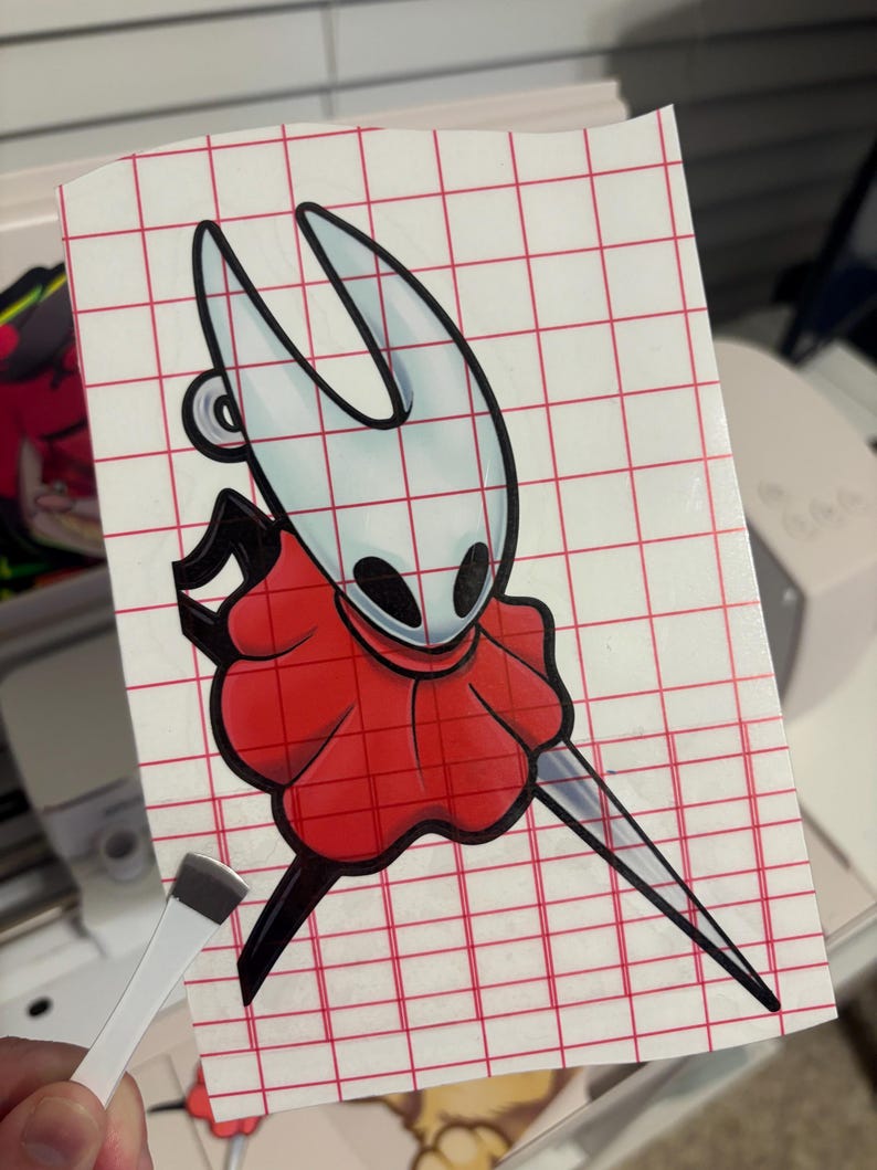 May include: A vinyl sticker featuring a cartoon character with a white head, black eyes, and a red cape. The character is holding a long, silver blade. The sticker is on a red grid background.