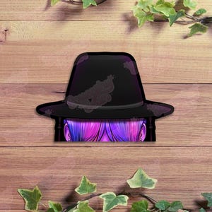 May include: A black fedora hat with a decorative design, partially obscuring a glimpse of purple and pink hair. The hat is set against a wooden background, with green leaves in the corners.