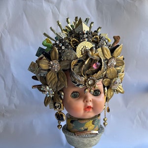 May include: A doll's head with a crown made of gold and jewel-toned trinkets. The crown is adorned with flowers and other decorative elements. The doll's head is on a gray pedestal.