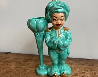May include: A turquoise ceramic figurine of a person wearing a turban and a long robe. The figurine has a gold trim and is holding a matching turquoise vase.