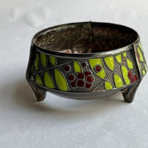 May include: A small, silver, footed bowl with a green and red enamel design. The bowl has a decorative pattern around the rim and three small feet.