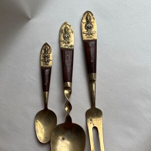 May include: A set of three vintage brass utensils with wooden handles. The utensils feature a spoon, a fork, and a serving spoon. Each handle has a gold Buddha figure and the word "SIAM" engraved on it.