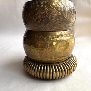 May include: Three brass bangles stacked on top of each other. The top bangle has an etched floral design. The middle bangle has a hammered texture, and the bottom bangle has a ribbed design. The bangles are a warm gold color.