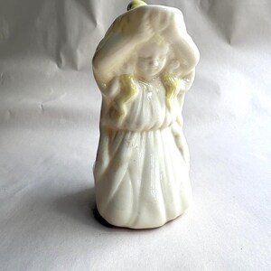 May include: A vintage ceramic figurine of a girl in a long white dress, with a large hat. The figurine has yellow accents in the hair and on the hat. The girl's head is bowed, and her hands are near her face.