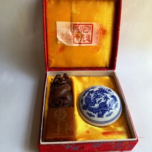 May include: A red box with a yellow satin lining contains two Chinese dragon seals. One seal is made of brown stone and the other is made of white porcelain with blue dragon designs. The box has a label that reads "KENNEDY"