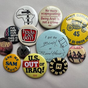 May include: A collection of vintage pinback buttons with various designs and text. The buttons feature phrases like "Texas Can Do It," "U.S. Out of Iraq," and "I got the Blues!" in a variety of colors and fonts.