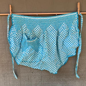 May include: A blue and white gingham apron with a ruffled edge and a single pocket. The apron is hanging on a wooden rod with clothespins.