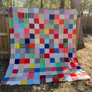 May include: A colorful patchwork quilt with a grid pattern of squares in various colors and patterns. The quilt is draped over a wooden fence and tree, showcasing a mix of red, blue, green, yellow, and patterned fabrics.