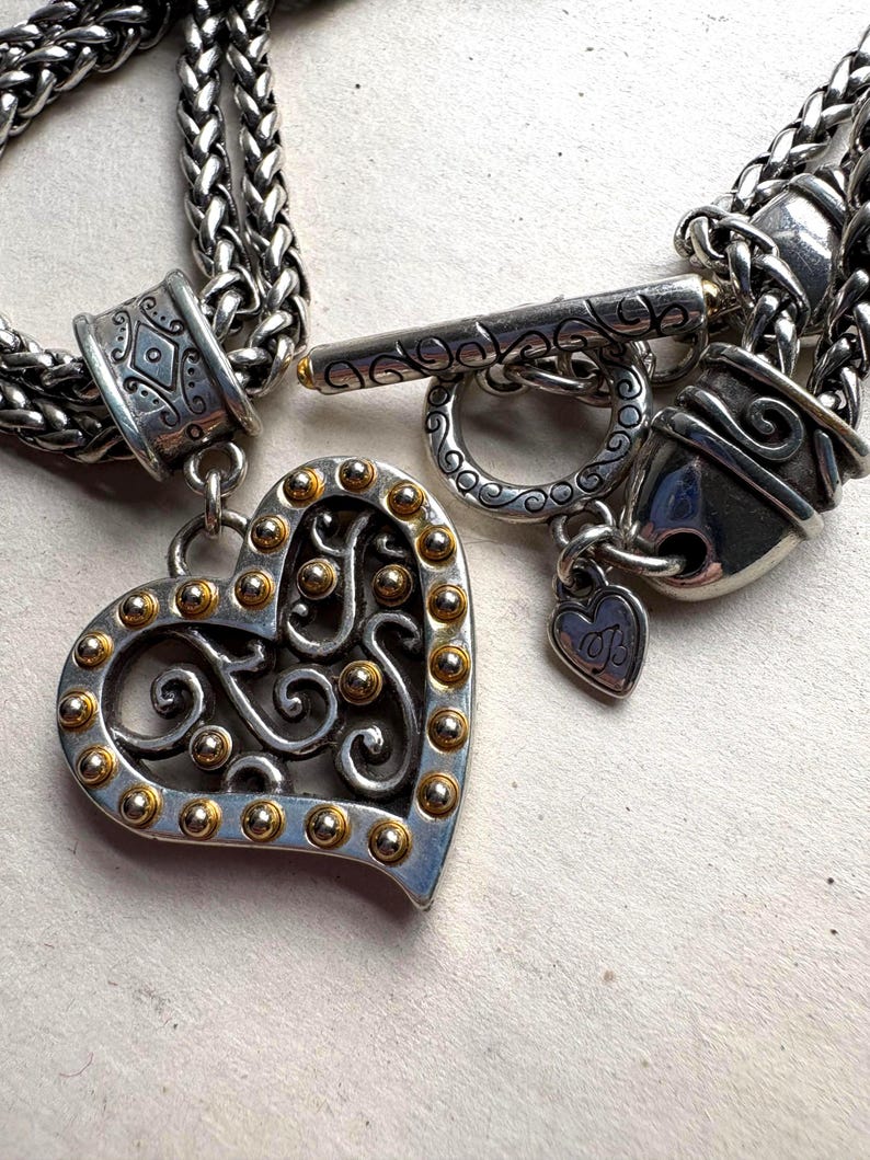 Pu&ograve; includere: Collana color argento con un ciondolo a forma di cuore. Il ciondolo presenta una cornice argentata con accenti dorati e un design traforato. La collana ha una chiusura a toggle e un piccolo ciondolo a forma di cuore. La catena &egrave; a treccia.