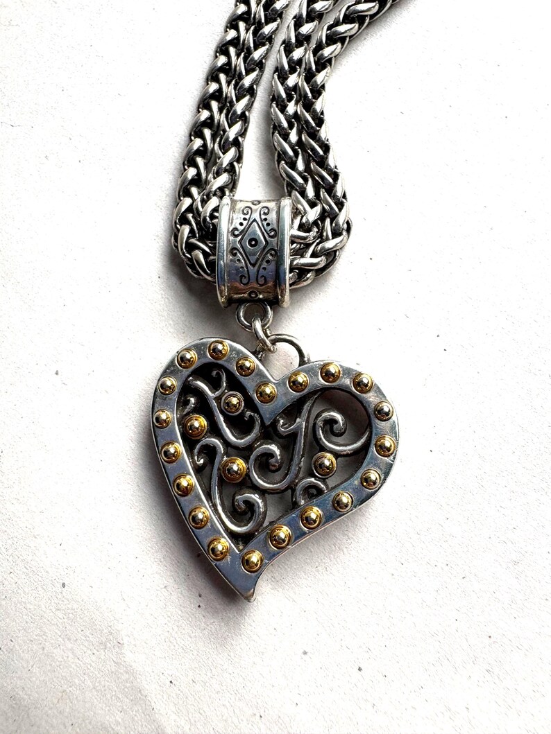 Pu&ograve; includere: Collana con pendente a forma di cuore argentato e dettagli dorati. Il cuore presenta un design traforato ed &egrave; impreziosito da piccole sfere dorate sui bordi. La catena &egrave; argentata.