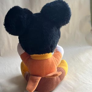 Op de afbeelding: Een pluche Mickey Mouse-speelgoed, van achteren bekeken. Het speelgoed heeft grote zwarte oren, een oranje shirt, bruine broek en gele schoenen. Het speelgoed zit, met de armen om de knie&euml;n.