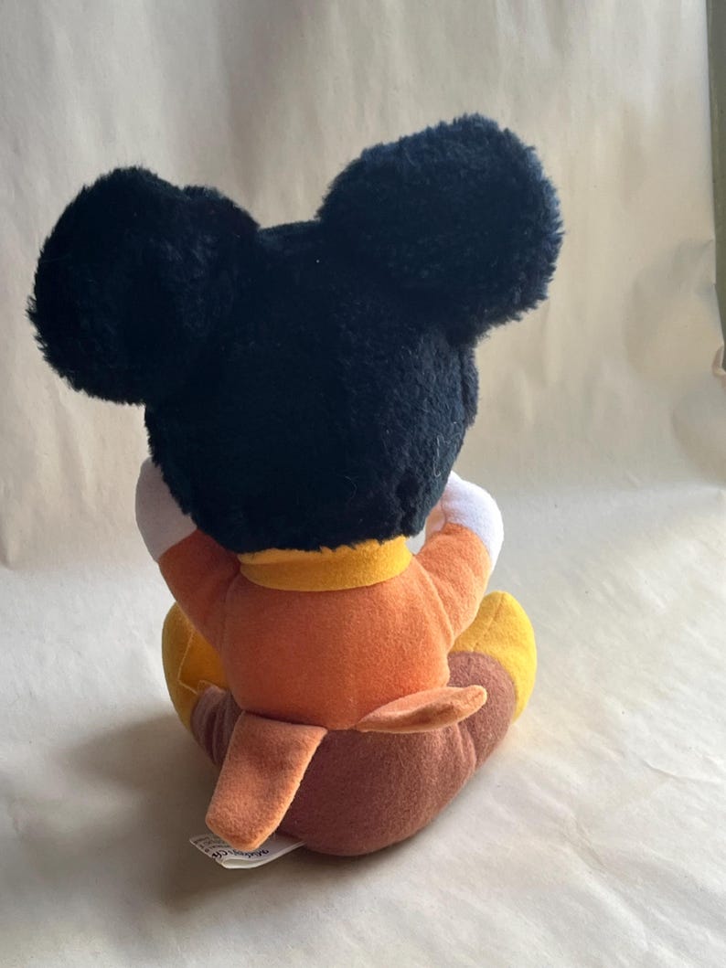 Op de afbeelding: Een pluche Mickey Mouse-speelgoed, van achteren bekeken. Het speelgoed heeft grote zwarte oren, een oranje shirt, bruine broek en gele schoenen. Het speelgoed zit, met de armen om de knie&euml;n.