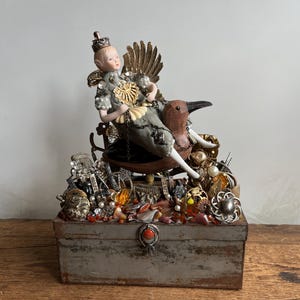 May include: A decorative box with a seated figurine riding a bird, adorned with a crown and gold accents. The box is overflowing with vintage jewelry, including pearls, brooches, and beads. The box has a metal clasp.