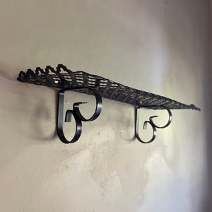 May include: Black metal wall shelf with a decorative, woven design. The shelf has two scrolled brackets.