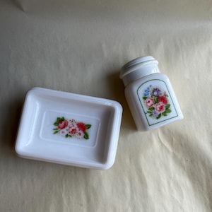 May include: A white soap dish and a small jar with a lid. Both items feature a floral design with red, pink, and blue flowers. The soap dish is rectangular, and the jar has a rounded shape with a green border around the floral design.