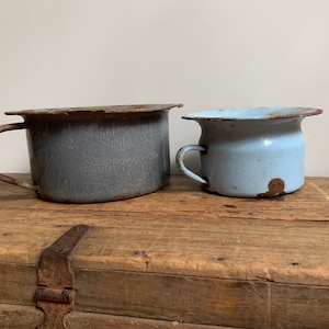 May include: Two vintage enamel chamber pots, one in light blue and one in gray, with rust and wear, sitting on a wooden surface.