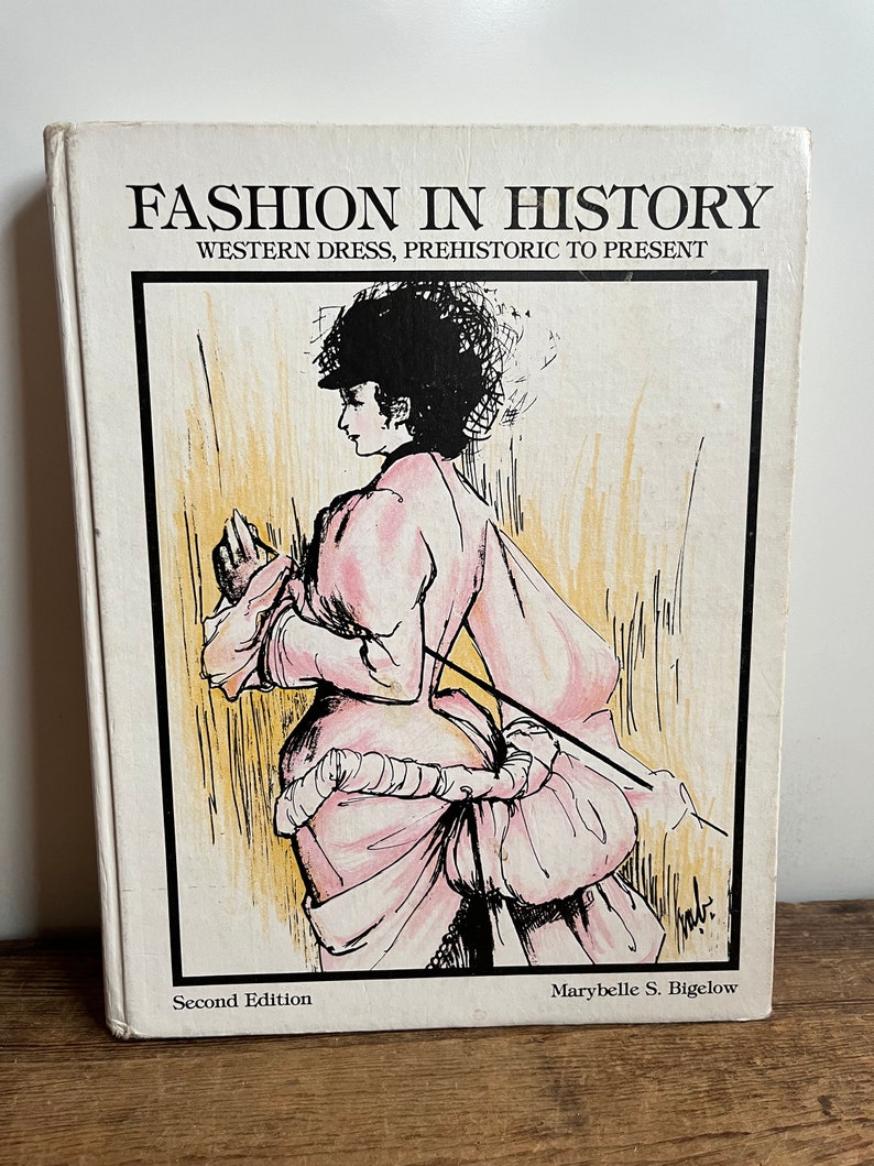May include: A book titled "Fashion in History: Western Dress, Prehistoric to Present" with a second edition copyright. The cover features a black and white illustration of a woman wearing a pink dress with a large bustle.