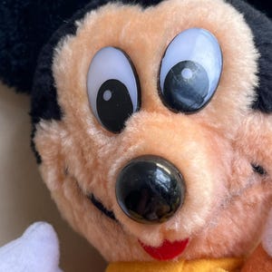 Op de afbeelding: Close-up van een vintage Mickey Mouse pluche speelgoed. Het pluche heeft grote, expressieve ogen, een zwarte neus en een rode mond. Het heeft zwarte oren, perzik-kleurig haar en een gele sjaal. Een klassiek Disney-personage.