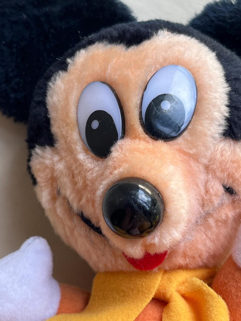 Op de afbeelding: Close-up van een vintage Mickey Mouse pluche speelgoed. Het pluche heeft grote, expressieve ogen, een zwarte neus en een rode mond. Het heeft zwarte oren, perzik-kleurig haar en een gele sjaal. Een klassiek Disney-personage.