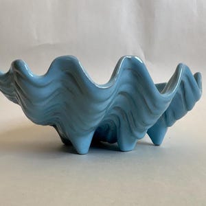 May include: A light blue ceramic bowl shaped like a giant clam shell. The bowl features a glossy finish with deep, undulating ridges and a scalloped edge that mimics the natural form of a mollusk shell, resting on three small, integrated feet.