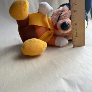 Op de afbeelding: Een vintage pluche speelgoed van een stripfiguur met een zwarte kop, grote zwarte ogen en een rode neus. Het speelgoed draagt een geel en oranje outfit met witte handschoenen en gele schoenen. Een houten liniaal is naast het speelgoed geplaatst.