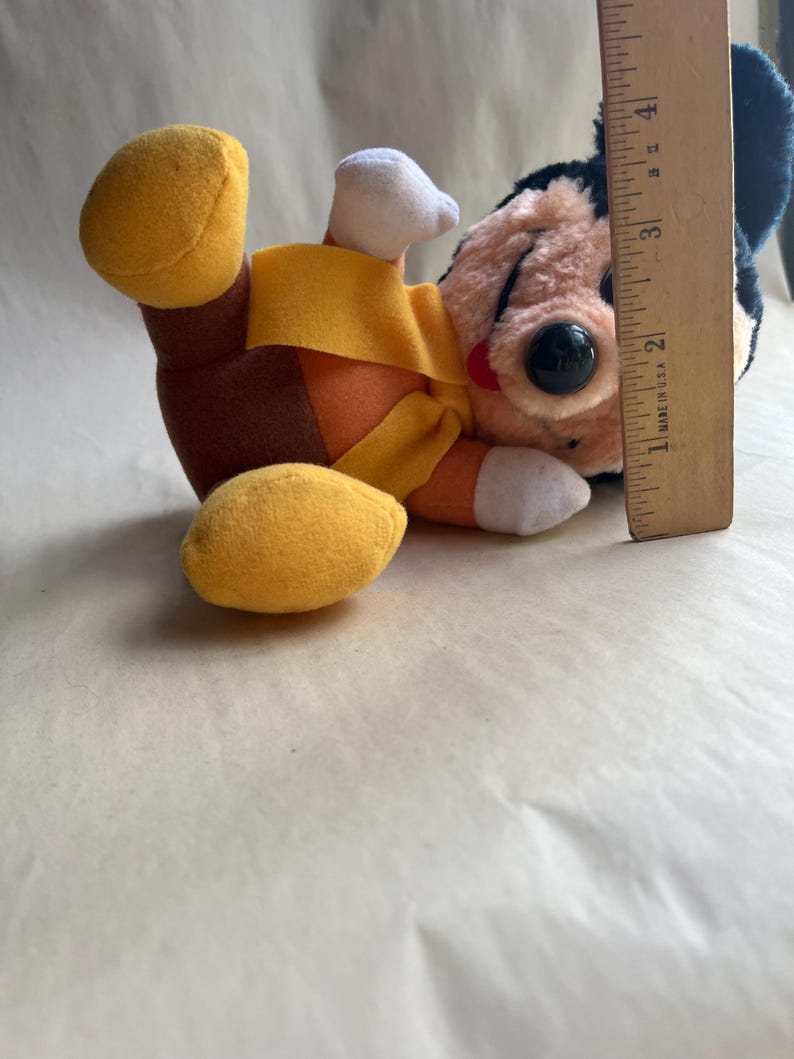 Op de afbeelding: Een vintage pluche speelgoed van een stripfiguur met een zwarte kop, grote zwarte ogen en een rode neus. Het speelgoed draagt een geel en oranje outfit met witte handschoenen en gele schoenen. Een houten liniaal is naast het speelgoed geplaatst.