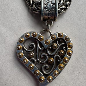 Pu&ograve; includere: Collana con pendente a cuore color argento con dettagli dorati. Il pendente a forma di cuore presenta un design traforato e presenta piccole borchie dorate sui bordi. La collana ha una spessa catena d'argento intrecciata.
