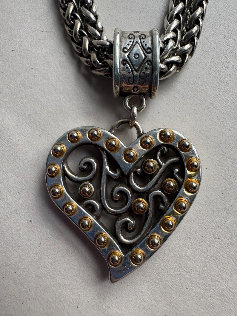 Pu&ograve; includere: Collana con pendente a cuore color argento con dettagli dorati. Il pendente a forma di cuore presenta un design traforato e presenta piccole borchie dorate sui bordi. La collana ha una spessa catena d'argento intrecciata.