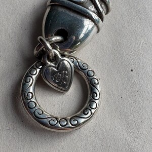 Pu&ograve; includere: Primo piano di un componente di braccialetto con ciondoli argentato. Il design presenta un ciondolo a forma di cuore con iniziali, un anello decorativo con volute e una chiusura strutturata. Il braccialetto &egrave; in metallo e ha un aspetto antico.