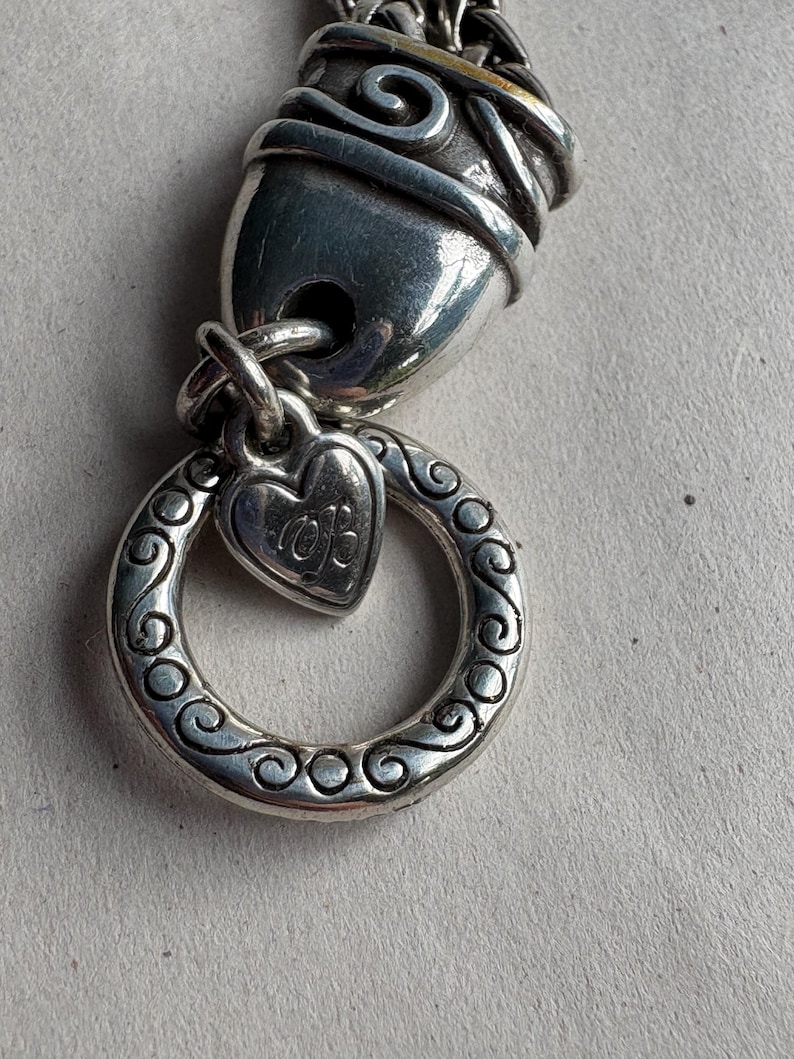 Pu&ograve; includere: Primo piano di un componente di braccialetto con ciondoli argentato. Il design presenta un ciondolo a forma di cuore con iniziali, un anello decorativo con volute e una chiusura strutturata. Il braccialetto &egrave; in metallo e ha un aspetto antico.