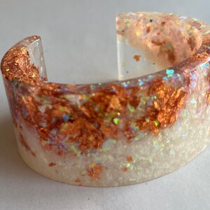 May include: A white resin bangle bracelet with copper and iridescent glitter embedded in the resin.