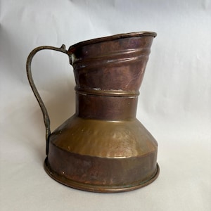 May include: A copper pitcher with a wide, flared base and a narrower, cylindrical upper section. The vessel features a curved metal handle attached to the side, with a distinct horizontal seam separating the two main body parts of the pitcher.