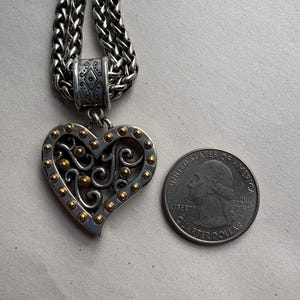 Pu&ograve; includere: Collana con pendente a forma di cuore color argento con dettagli dorati. Il pendente a forma di cuore presenta un design traforato ed &egrave; attaccato a una catena intrecciata. Una moneta da venticinque centesimi americana &egrave; accanto al pendente come riferimento delle dimensioni.
