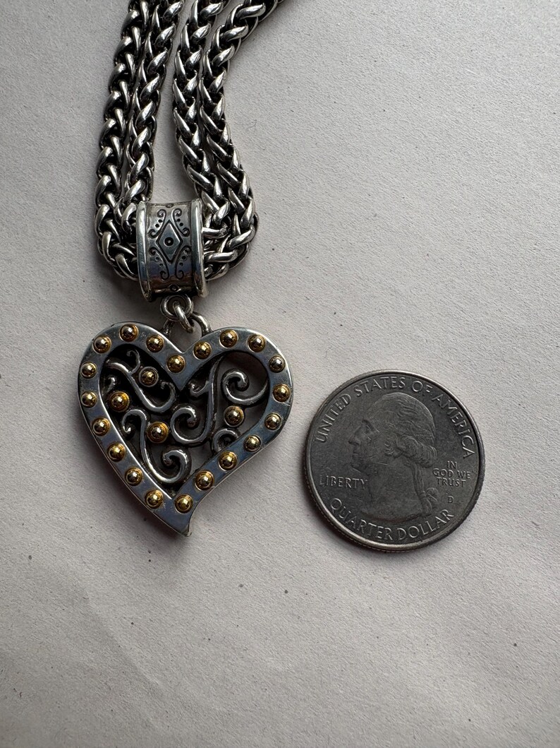 Pu&ograve; includere: Collana con pendente a forma di cuore color argento con dettagli dorati. Il pendente a forma di cuore presenta un design traforato ed &egrave; attaccato a una catena intrecciata. Una moneta da venticinque centesimi americana &egrave; accanto al pendente come riferimento delle dimensioni.