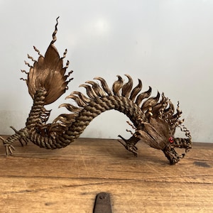 May include: A detailed, brown dragon sculpture with a textured, woven body and a large, fan-shaped tail. The dragon has a curved posture, with a red gem in its eye, and is displayed on a wooden surface.
