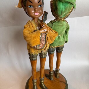 May include: Two elf figurines standing on a circular wooden base. One elf wears a mustard yellow velvet outfit and holds a small stringed instrument. The other elf wears a green velvet outfit. Both have pointed hats with gold trim and brown boots.