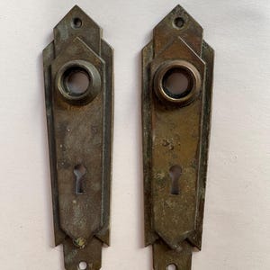 May include: A pair of weathered brass door plates featuring a geometric, elongated hexagonal shape with pointed ends. Each plate includes a circular knob opening at the top and a traditional keyhole cutout below, with screw holes at the top and bottom.