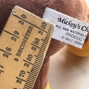 Op de afbeelding: Close-up van een vintage Mickey Mouse pluche speelgoed met een bruin lichaam en gele accenten. Een houten liniaal toont metingen in decimeters. Het label luidt "Mickey's Ch" en "ALL NEW MATERIALS PRODUCT &copy; WALT DISNEY".