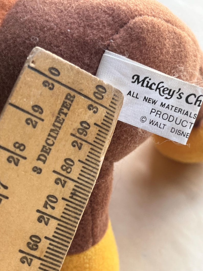 Op de afbeelding: Close-up van een vintage Mickey Mouse pluche speelgoed met een bruin lichaam en gele accenten. Een houten liniaal toont metingen in decimeters. Het label luidt "Mickey's Ch" en "ALL NEW MATERIALS PRODUCT &copy; WALT DISNEY".