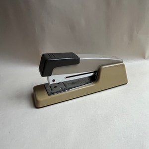 May include: A beige and gray metal stapler with a black top. The stapler has a clear plastic window to view the staples.