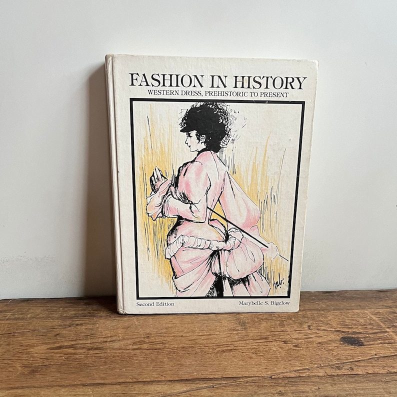 May include: A vintage book titled "Fashion in History: Western Dress, Prehistoric to Present" by Marybelle S. Bigelow. The book cover features a black and white illustration of a woman in a pink dress.