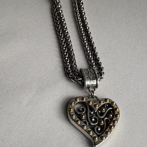Pu&ograve; includere: Collana color argento con pendente a forma di cuore. Il pendente presenta un bordo argentato con accenti dorati e un design a filigrana decorativo. La catena &egrave; in stile intrecciato.