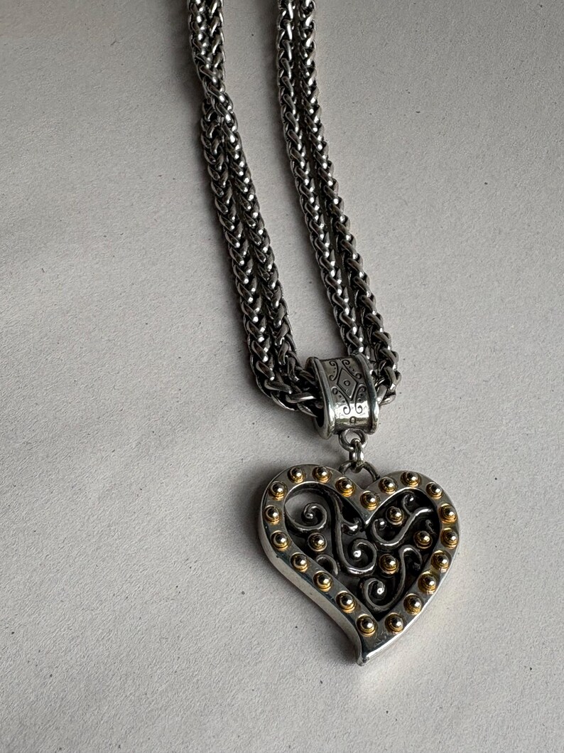 Pu&ograve; includere: Collana color argento con pendente a forma di cuore. Il pendente presenta un bordo argentato con accenti dorati e un design a filigrana decorativo. La catena &egrave; in stile intrecciato.
