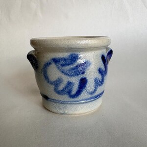 May include: A small, cylindrical stoneware crock with a light gray, textured surface and a thick, rounded rim. The vessel features two small side handles and is decorated with hand-painted cobalt blue floral patterns encircling the exterior.