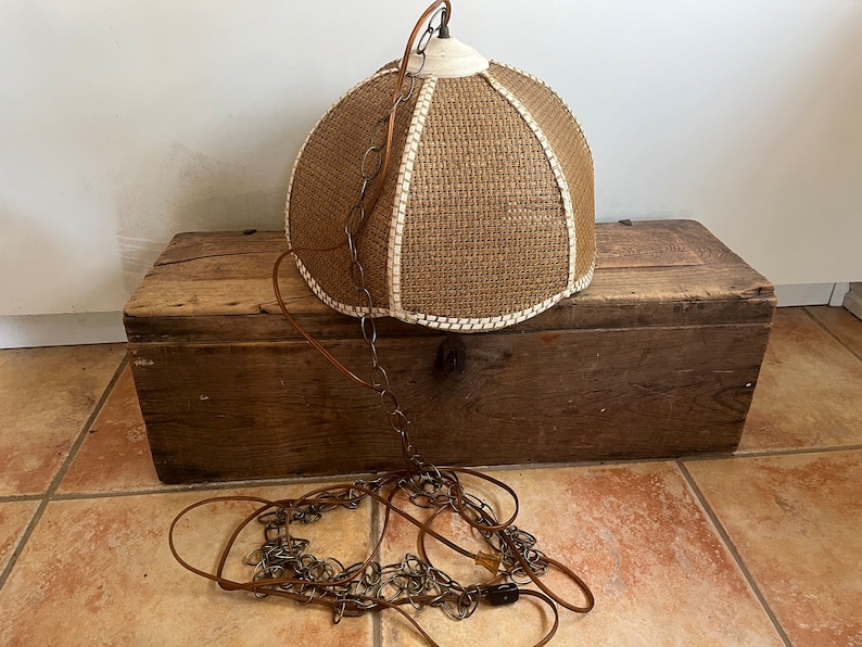 Puede incluir: Una l&aacute;mpara colgante de tejido marr&oacute;n con un borde blanco y una cadena que cuelga de ella. La l&aacute;mpara est&aacute; colocada encima de una caja de madera.
